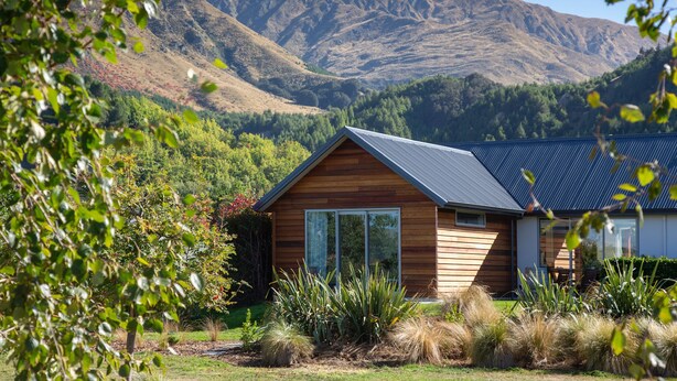 A modern house with a sloped roof, surrounded by a well-maintained garden and a mountainous backdrop. - The Arrow Nest - Beautiful Stylish Apartment (Arrowtown)