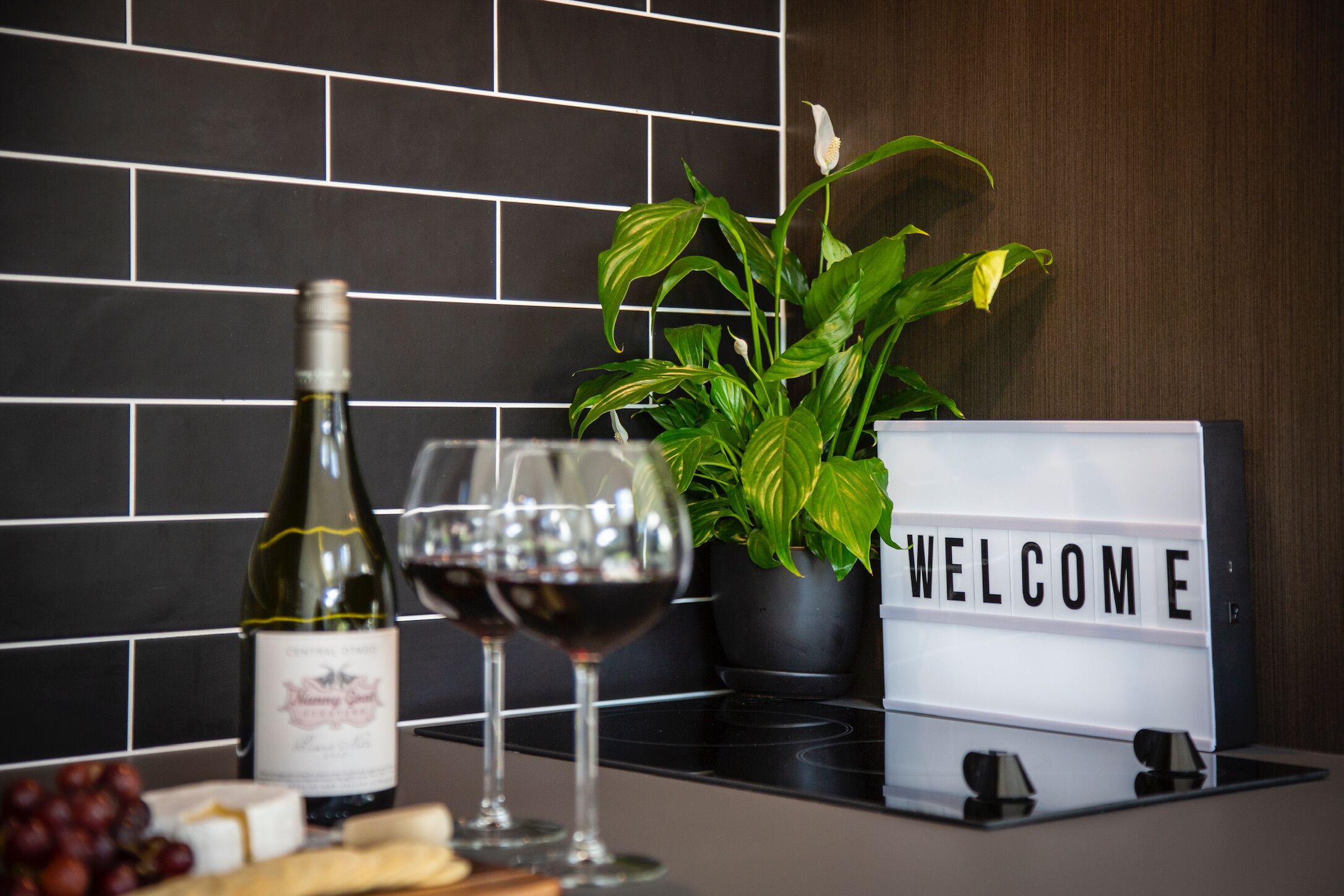 A kitchen counter with a wine bottle, two wine glasses, a welcome sign, a potted plant, and some cheese and grapes.