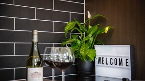 A kitchen counter with a wine bottle, two wine glasses, a welcome sign, a potted plant, and some cheese and grapes. - The Arrow Nest - Beautiful Stylish Apartment (Arrowtown)
