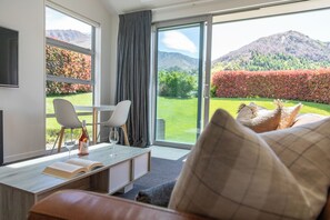 A modern hotel room with a large window, a sofa, a table with a book and wine bottle, and a view of mountains and a garden. - The Arrow Nest - Beautiful Stylish Apartment (Arrowtown)