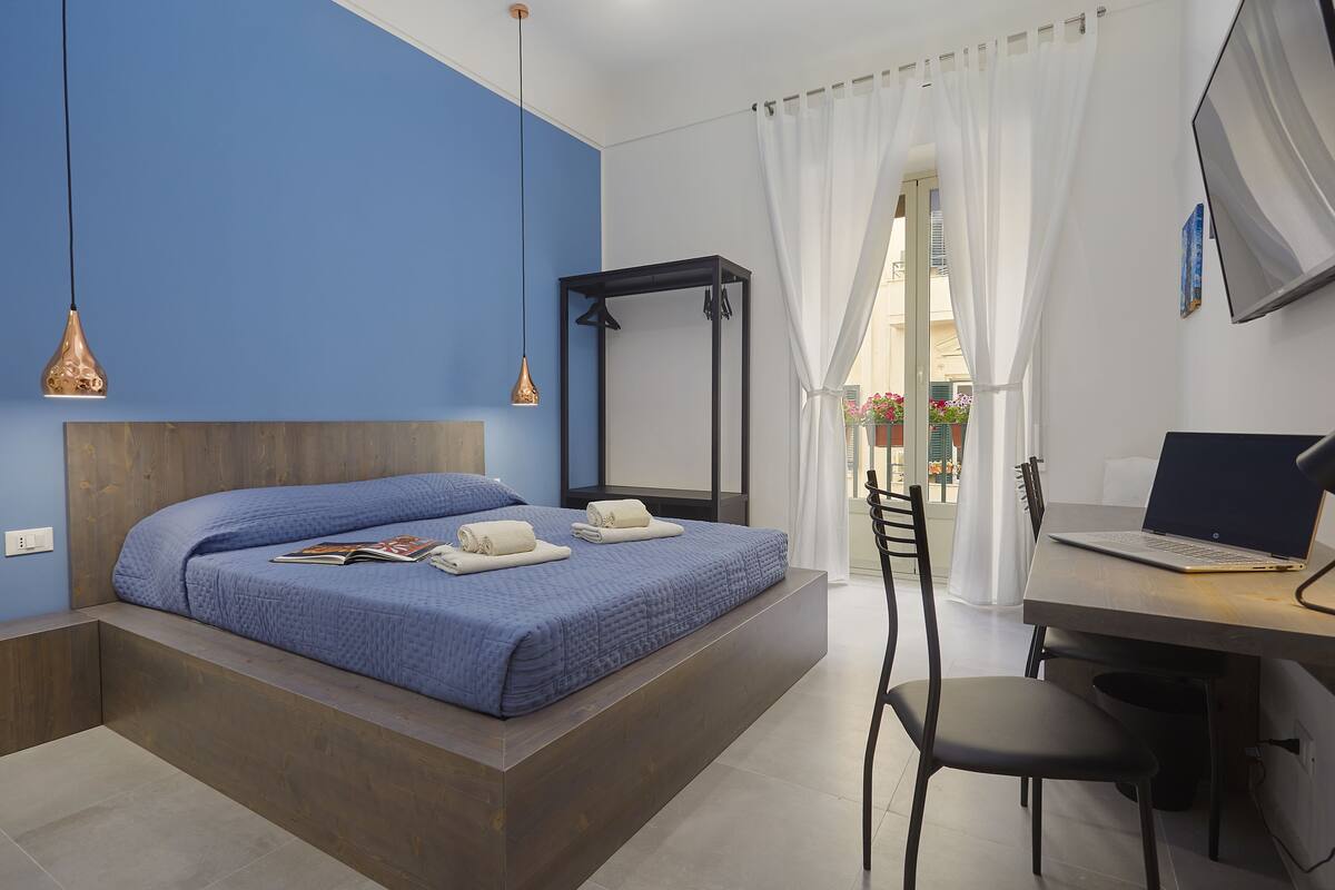 A bedroom with a bed, a desk with a laptop, and a window with curtains.