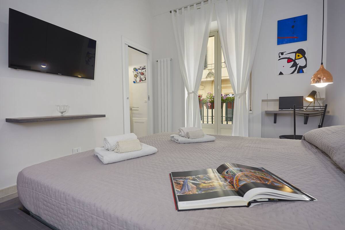 A modern hotel room with a large bed, a flat-screen TV, a laptop on a desk, and a magazine on the bed.