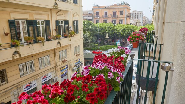 A balcony with red and pink flowers, a view of a street with cars, and buildings with balconies. - Better & Better (Palermo)