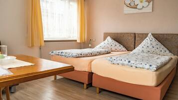 A room with two beds, a wooden table, a window with yellow curtains, and a wall art piece.