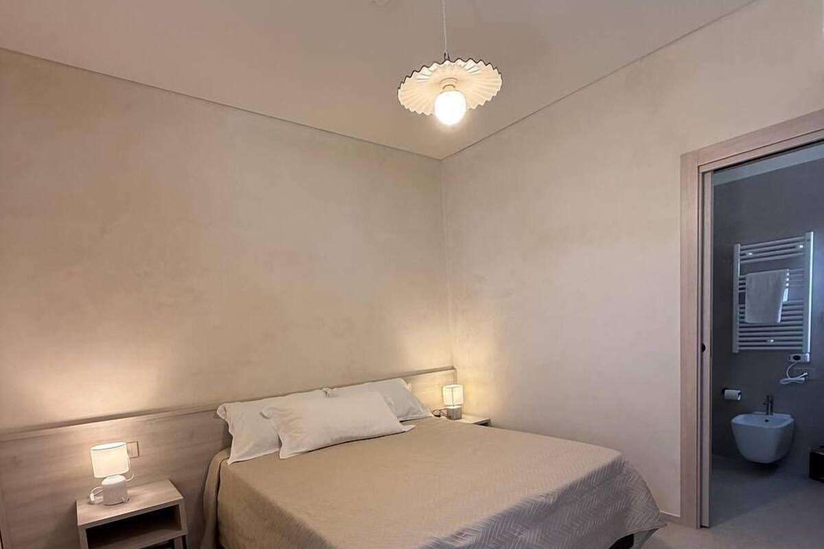 Et soveværelse med seng, sengeborde, et natbord, en vægmonteret lampe og et badeværelse med bidét og håndklædestang.
