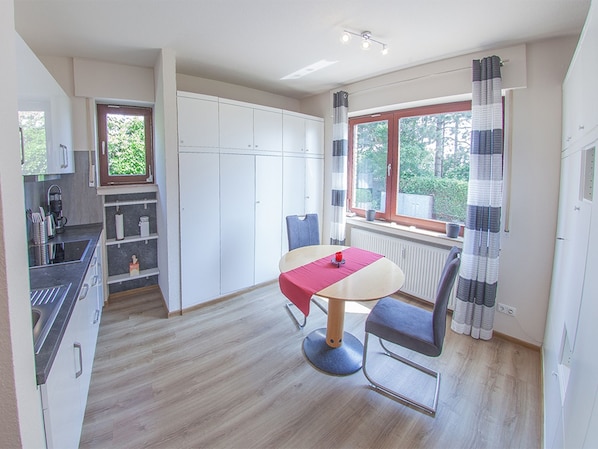 Eine kleine Küche mit modernen Geräten, ein Essbereich mit Tisch und Stühlen sowie ein Fenster mit Vorhängen.