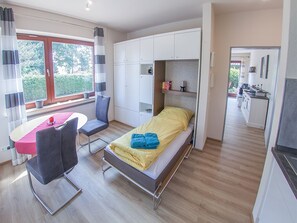 A compact room with a bed, a desk, and a chair. There is a window with curtains, a kitchenette, and a view of greenery outside.