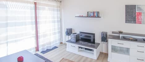 A modern living room with a flat-screen TV, a coffee table, a sofa, a dining table, and a kitchen area with a countertop and cabinets.