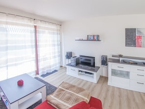 A modern living room with a flat-screen TV, a coffee table, a sofa, a dining table, and a kitchen area with a countertop and cabinets.