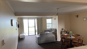 A living room with a grey sofa, a dining table with chairs, a ceiling fan, and a sliding glass door leading to a balcony. - 'Horizons' Unit 2 - Beachfront Unit (BARGARA)