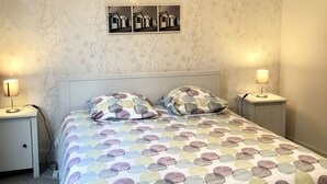 Un lit bien fait, recouvert d’une couvre-lit aux motifs circulaires et colorés, deux lampes de chevet, un tableau représentant des maisons de plage accroché au mur et un luminaire suspendu.