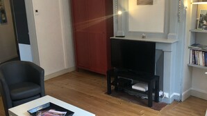 Un salon avec une table basse blanche, un fauteuil noir, une armoire rouge, un meuble TV noir et une cheminée surmontée d’un tableau encadré.