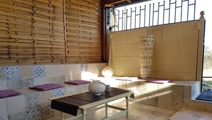 Um quarto com parede de madeira, piso de azulejos, um banco com almofadas roxas, uma mesa com tigela decorativa e porta estampada.