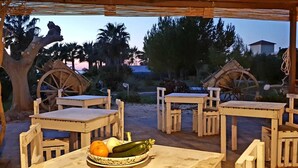 A rustic outdoor seating area with wooden tables and chairs, surrounded by palm trees and a large wooden wheel structure. - MammaColette (Marsala)