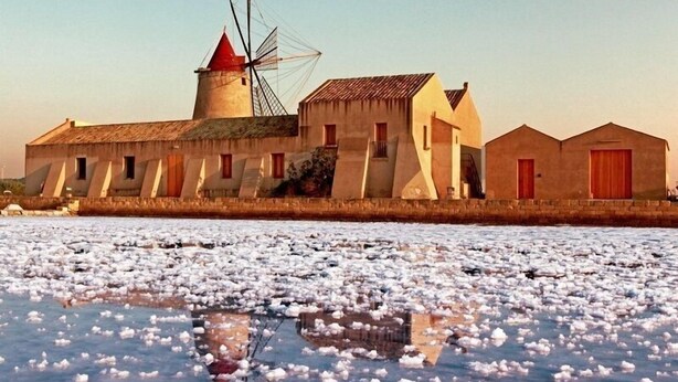 A coastal building with a windmill, a water body with floating objects, and a clear sky. - MammaColette (Marsala)