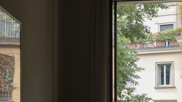 Une chambre d’hôtel avec un lit, une petite table avec une bouilloire, une poubelle et une vue sur un balcon doté d’une balustrade et agrémenté de quelques plantes.