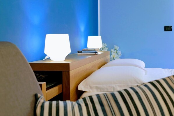 Un dormitorio con una mesita de noche de madera, dos lámparas de noche, una pila de libros y una cama con almohadas a rayas.