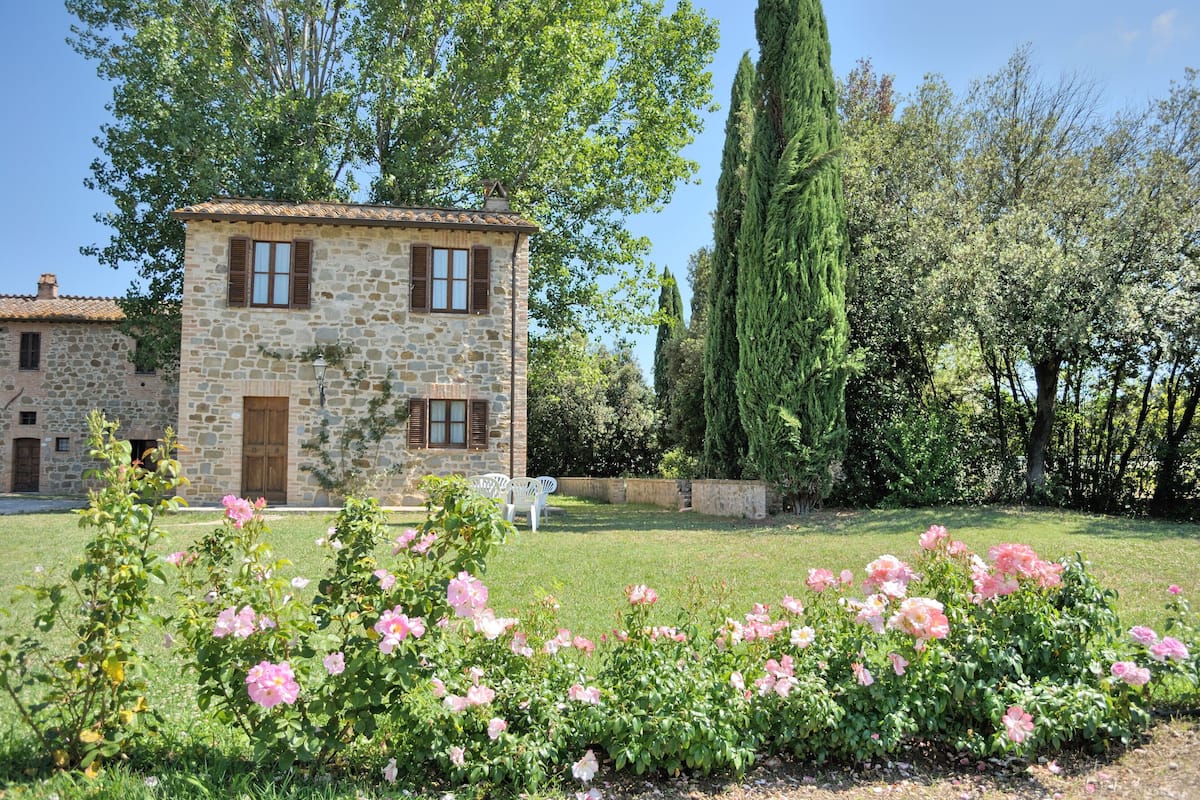 A stone building with a tiled roof, surrounded by a well-maintained garden with pink flowers.