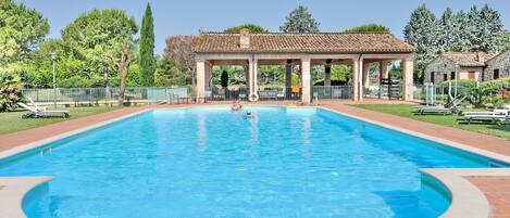 Une piscine rectangulaire entourée d’une terrasse carrelée, un pavillon couvert avec des colonnes et une clôture délimitant l’espace.
