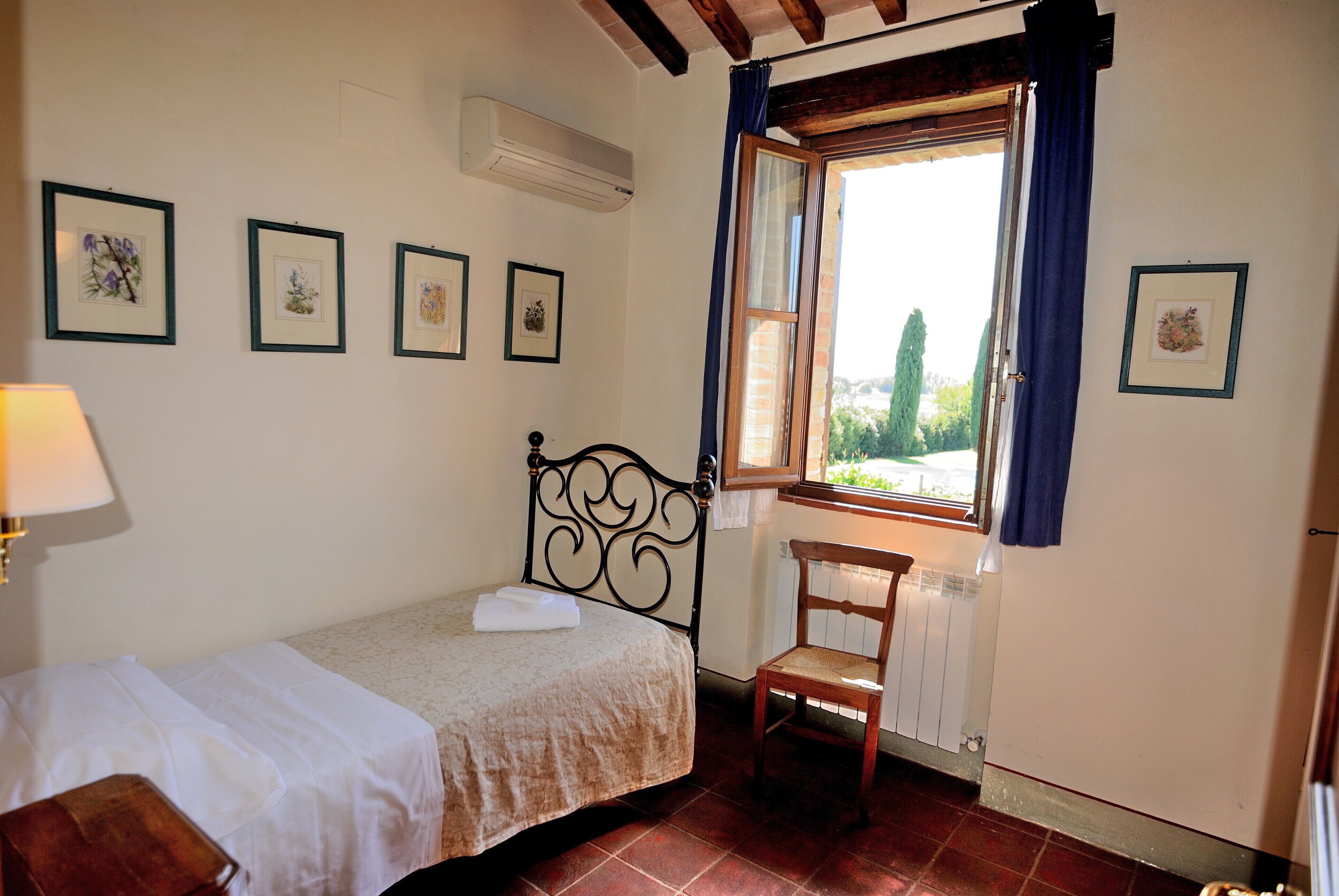 Une petite pièce propre et bien rangée, avec un lit, une chaise et une fenêtre avec des rideaux. Des tableaux encadrés ornent le mur et un appareil de climatisation est installé au-dessus du lit.