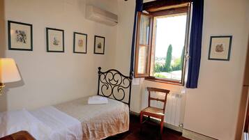 Une petite pièce propre et bien rangée, avec un lit, une chaise et une fenêtre avec des rideaux. Des tableaux encadrés ornent le mur et un appareil de climatisation est installé au-dessus du lit.
