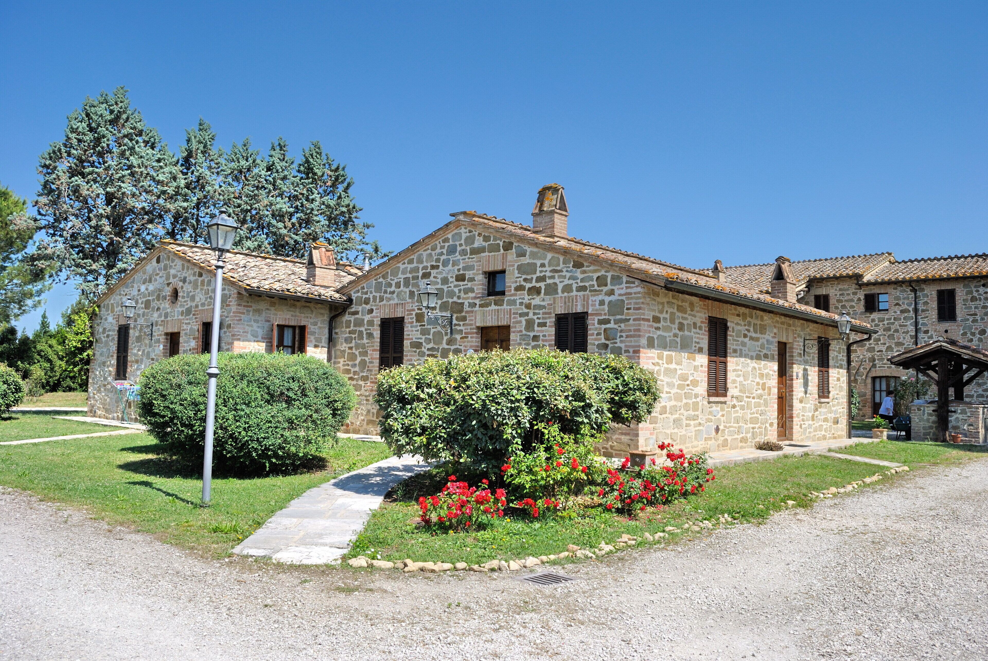 A stone building with a tiled roof, a small garden with red flowers, and a clear blue sky.