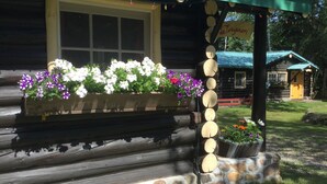 A log cabin with a porch, flower boxes with white and purple flowers, and a stone wall. - Domaine le Bostonnais (La Bostonnais)