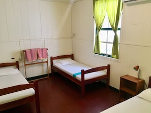 A room with two wooden beds, a window with green curtains, a bedside table with a lamp, and a wall-mounted telephone. - Guesthouse De Kleine Historie (Paramaribo)