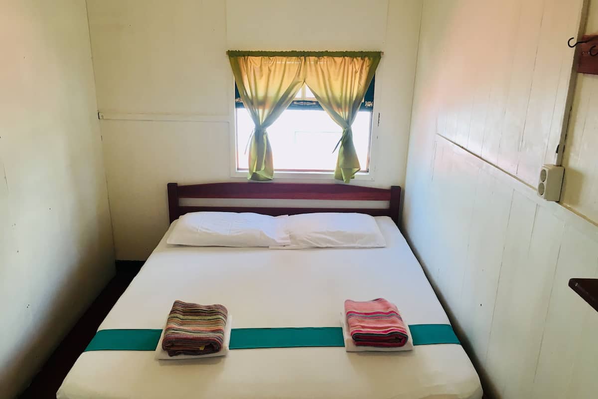 Un lit simple avec un matelas blanc, deux oreillers et une couverture verte. Il y a une fenêtre avec un rideau à moitié tiré, et un petit haut-parleur fixé au mur, sur la droite.