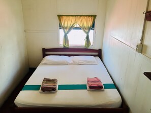 A single bed with a white mattress, two pillows, and a green bedspread. There is a window with a curtain partially drawn, and a small speaker mounted on the wall to the right. - Guesthouse De Kleine Historie (Paramaribo)