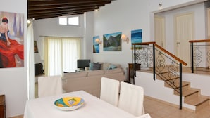 A living room with a sofa, a dining table with a plate, a staircase with a railing, and wall art. - Orestis Suites (Skiathos)