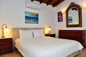 Cama bem arrumada com lençóis brancos, mesa de cabeceira de madeira com abajur, cômoda com gavetas e duas luminárias fixadas na parede.