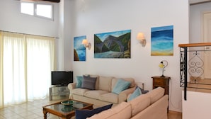 A living room with a sofa, a coffee table, and a TV. There are paintings on the wall and a window with curtains. - Orestis Suites (Skiathos)