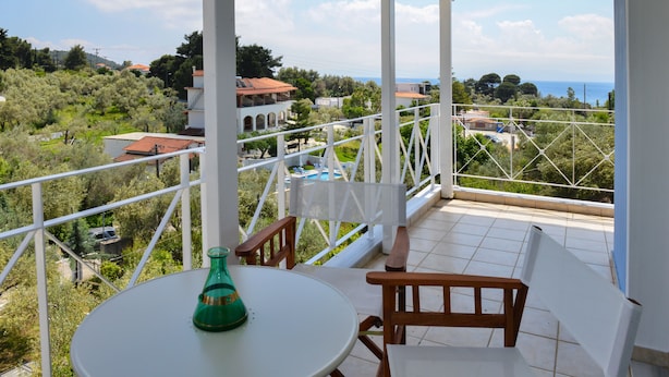 A balcony with a white table and chairs, overlooking a scenic view of a coastal area with houses and greenery. - Orestis Suites (Skiathos)