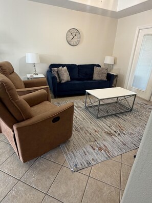 A living room with a brown armchair, a blue sofa, a white coffee table, and a wall clock.