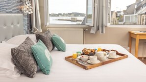 Een hotelkamer met een bed, een ontbijtplateau, kussens en uitzicht op het water en gebouwen.