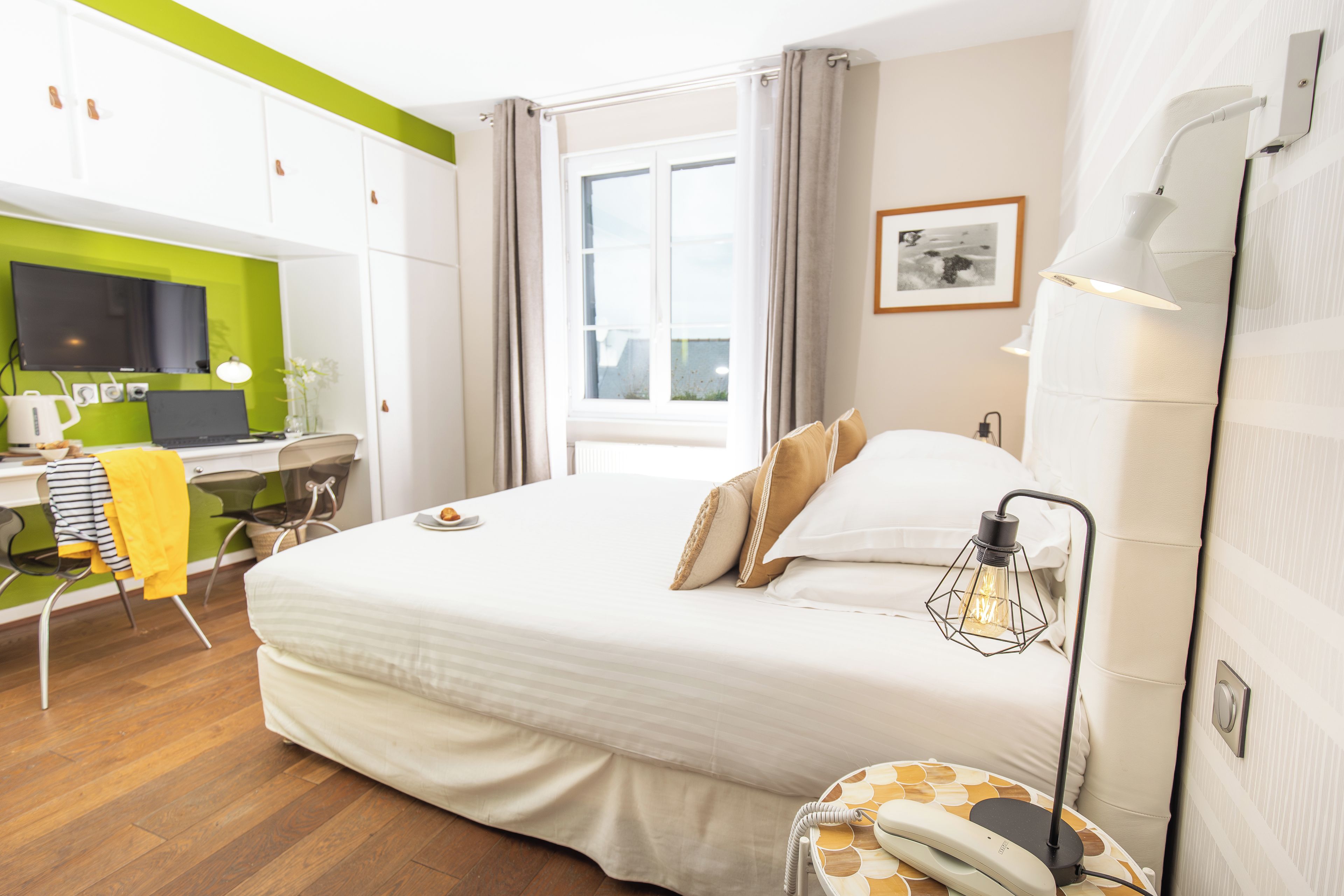 A hotel room with a bed, a desk with a laptop, a chair, a bedside table with a lamp, and a window.