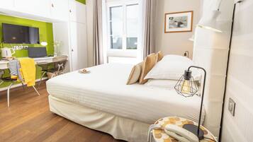 A hotel room with a bed, a desk with a laptop, a chair, a bedside table with a lamp, and a window.