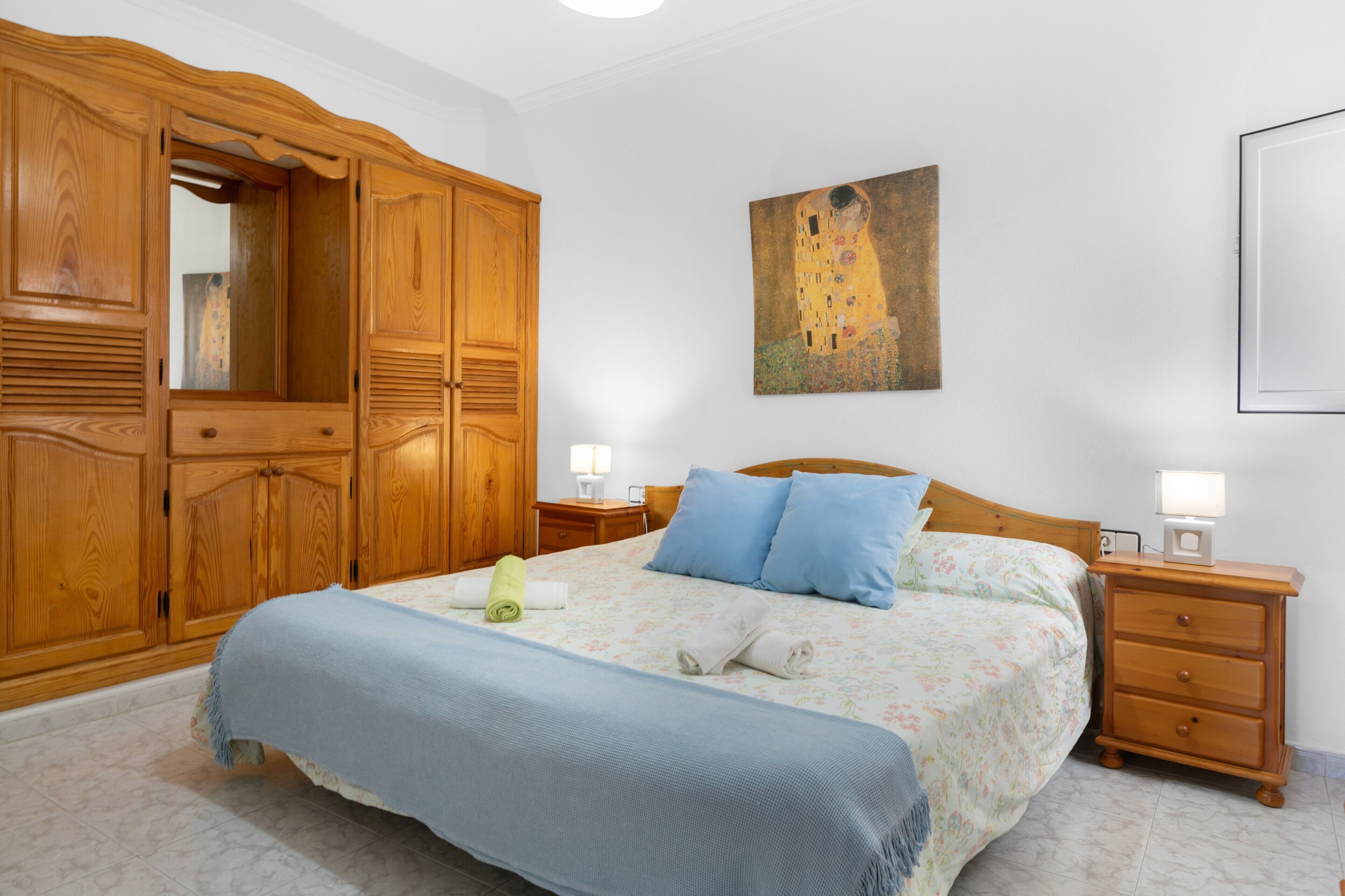 Un dormitorio con un armario grande de madera, una cama con ropa de cama azul y blanca, un cuadro en la pared y una mesita de noche con una lámpara.