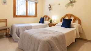 Une chambre avec deux lits simples, un banc, une fenêtre avec des stores, une lampe fixée au mur et un élément décoratif mural.