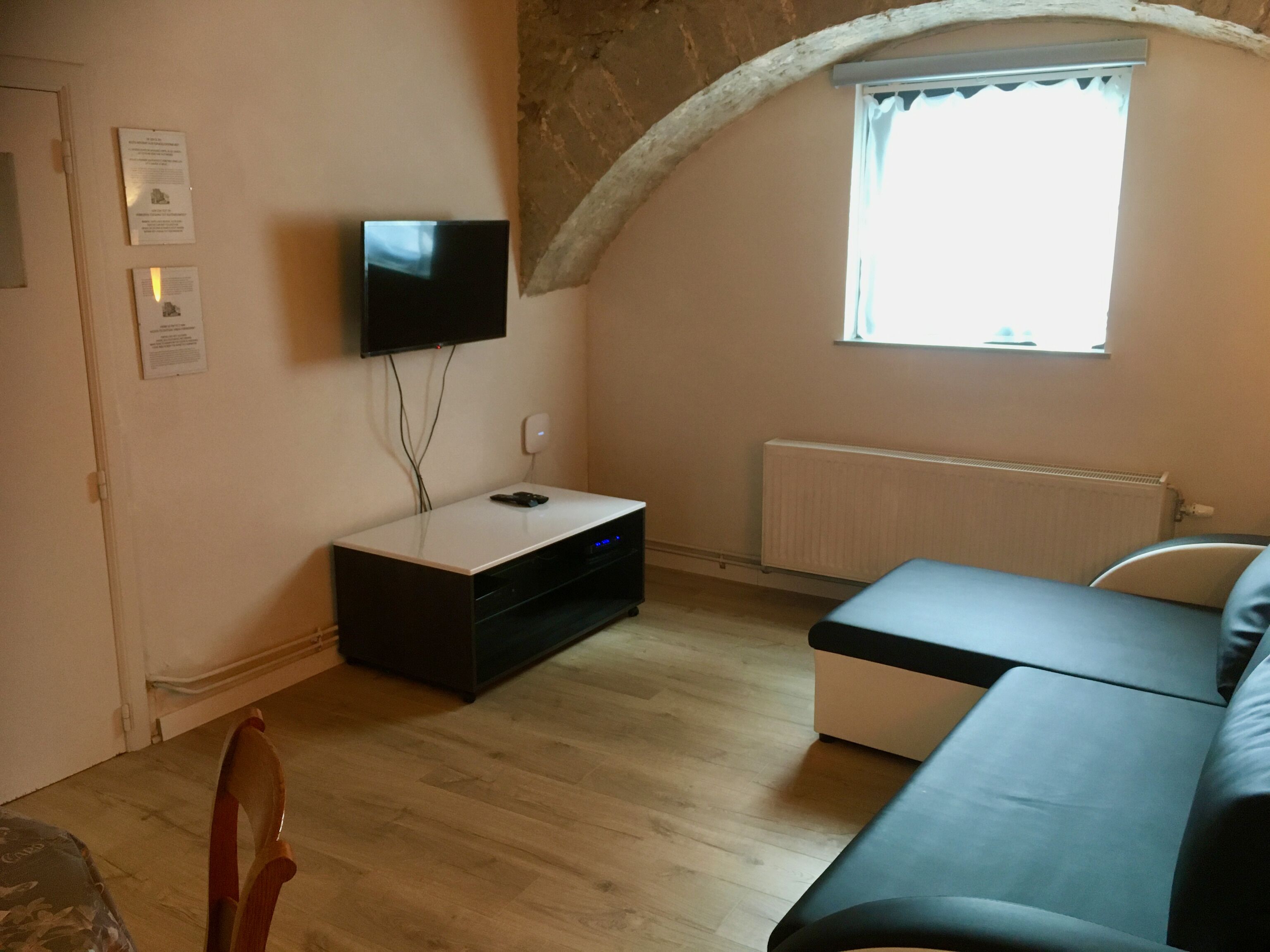 A small, cozy room with a flat-screen TV mounted on the wall, a wooden floor, a window with a view, and a bed with a headboard.