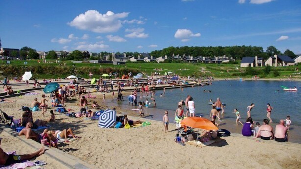A beach with people sunbathing and swimming, a pier extending into the water, and houses in the background. - Châtelet de Barbençon (Beaumont)