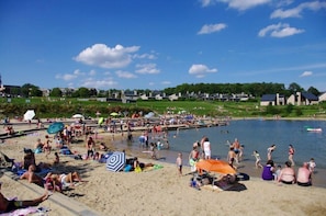 A beach with people sunbathing and swimming, a pier extending into the water, and houses in the background. - Châtelet de Barbençon (Beaumont)
