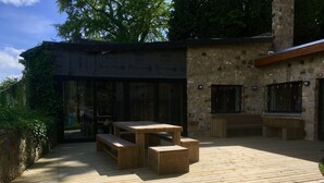 An outdoor patio area with a wooden table and benches, surrounded by stone walls and glass doors. - Châtelet de Barbençon (Beaumont)