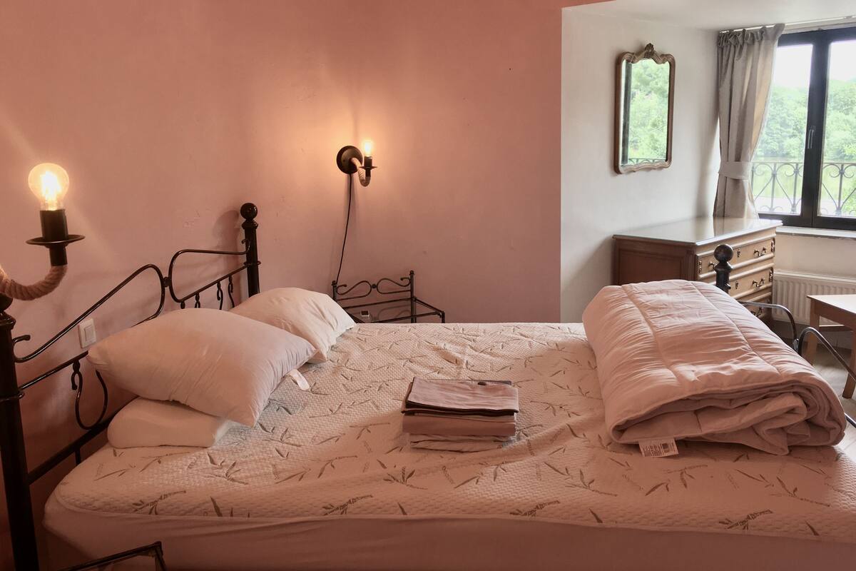 Una cama bien hecha con un edredón blanco y almohadas, una mesita de noche con una lámpara, y un aparador con un espejo.