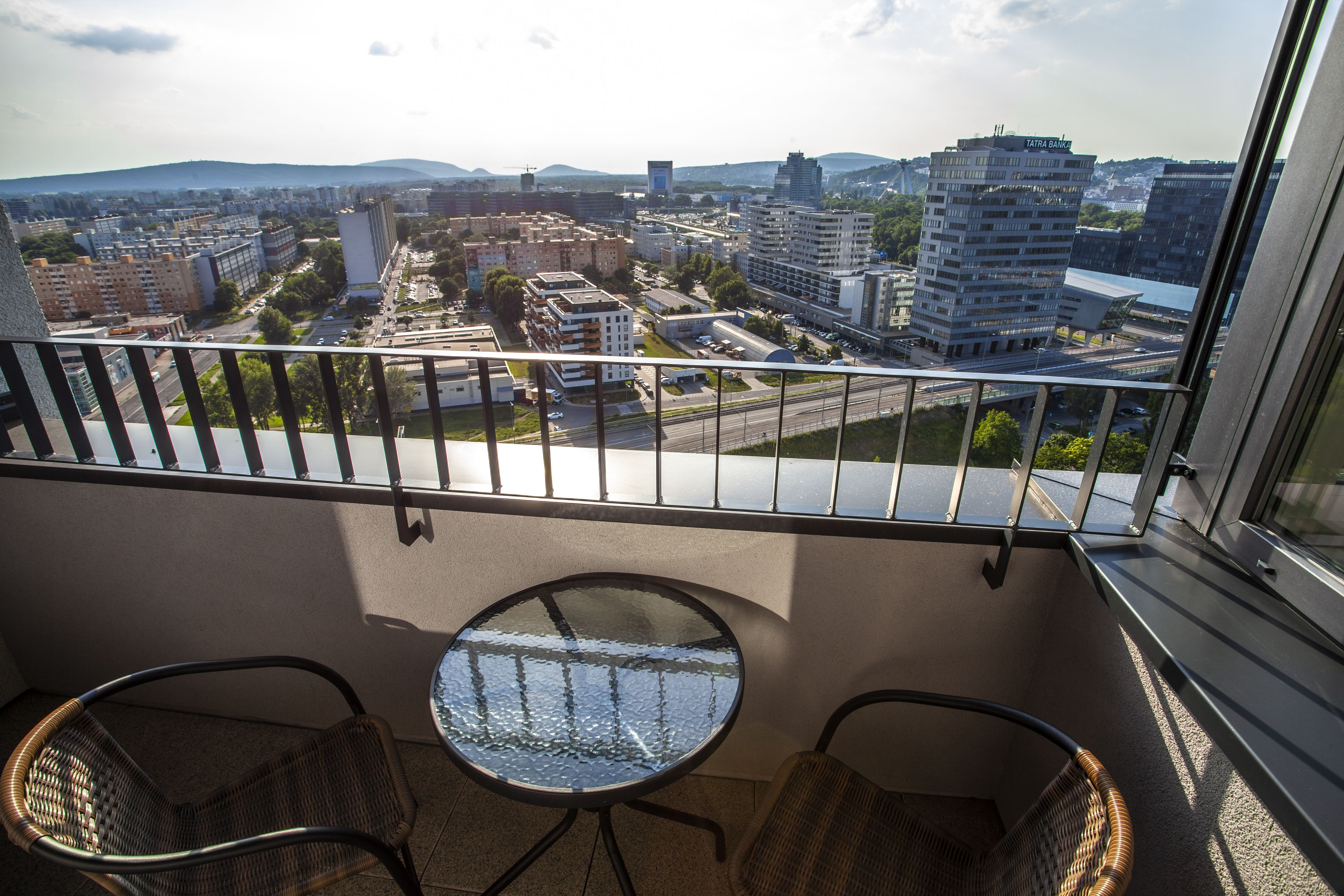 Een balkon met twee rieten stoelen en een bijzettafeltje, met uitzicht op een stadsgezicht met gebouwen en een snelweg.