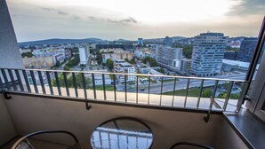 Ein Balkon mit Korbstühlen, einem kleinen runden Tisch und Blick auf eine Stadtansicht, darunter ein Bankgebäude.