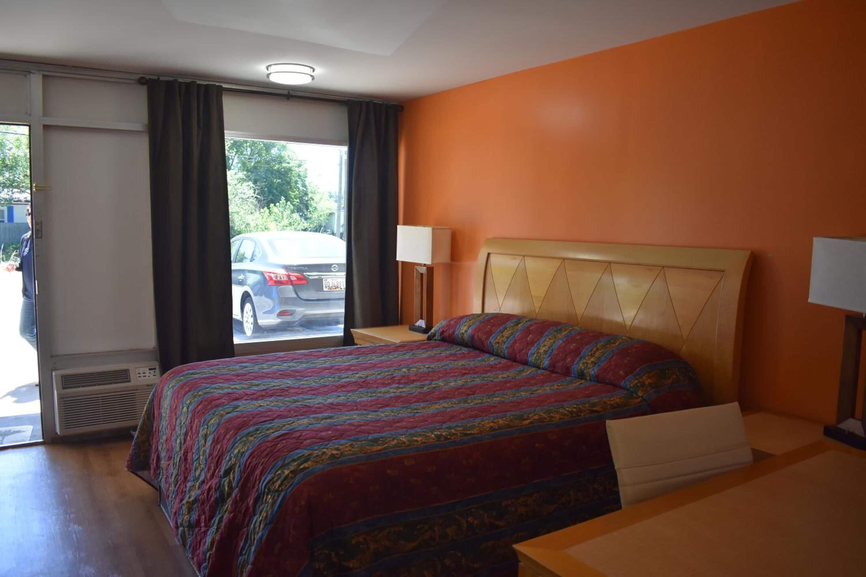 A hotel room with a bed, a bedside table, a lamp, and a view of the street through the window.