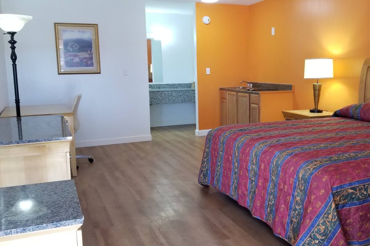 A hotel room with a bed, desk, chair, lamp, and a small kitchenette.