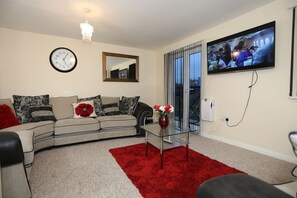 A living room with a sofa, a glass coffee table, a red rug, a clock, a mirror, and a TV. - Beautiful 4 beds Hillview House (London)